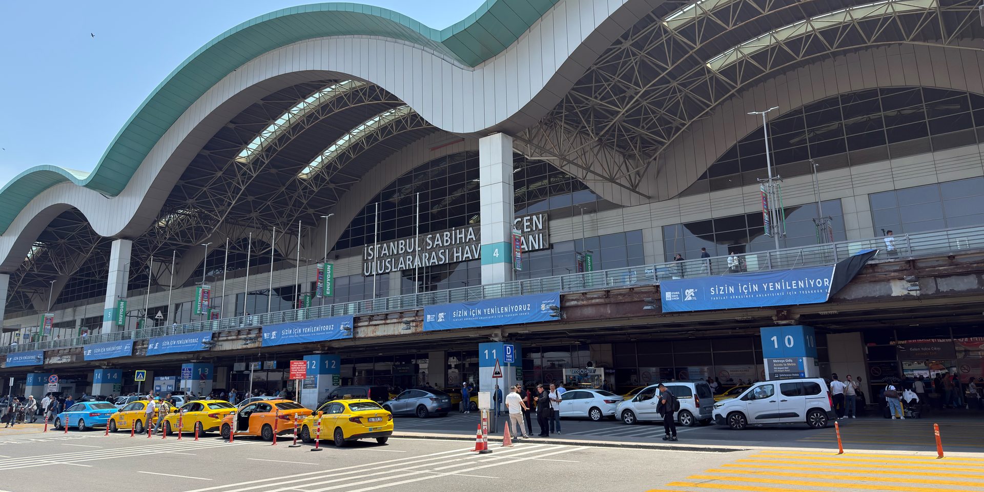 The Sabiha Gökçen airport terminal frontage with its wavy white roof, yellow taxis waiting at the kerb and a zebra crossing in front.