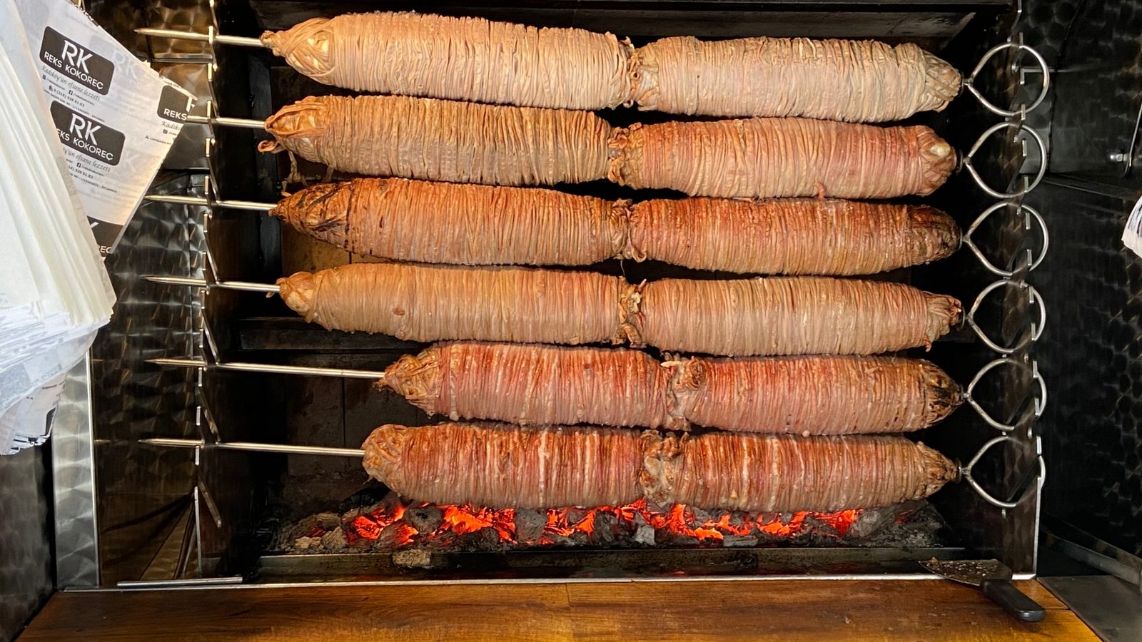 Six kokoreç rolls on stacked metal skewers grilling over charcoal embers in a stainless-steel cabinet at an Istanbul street stand