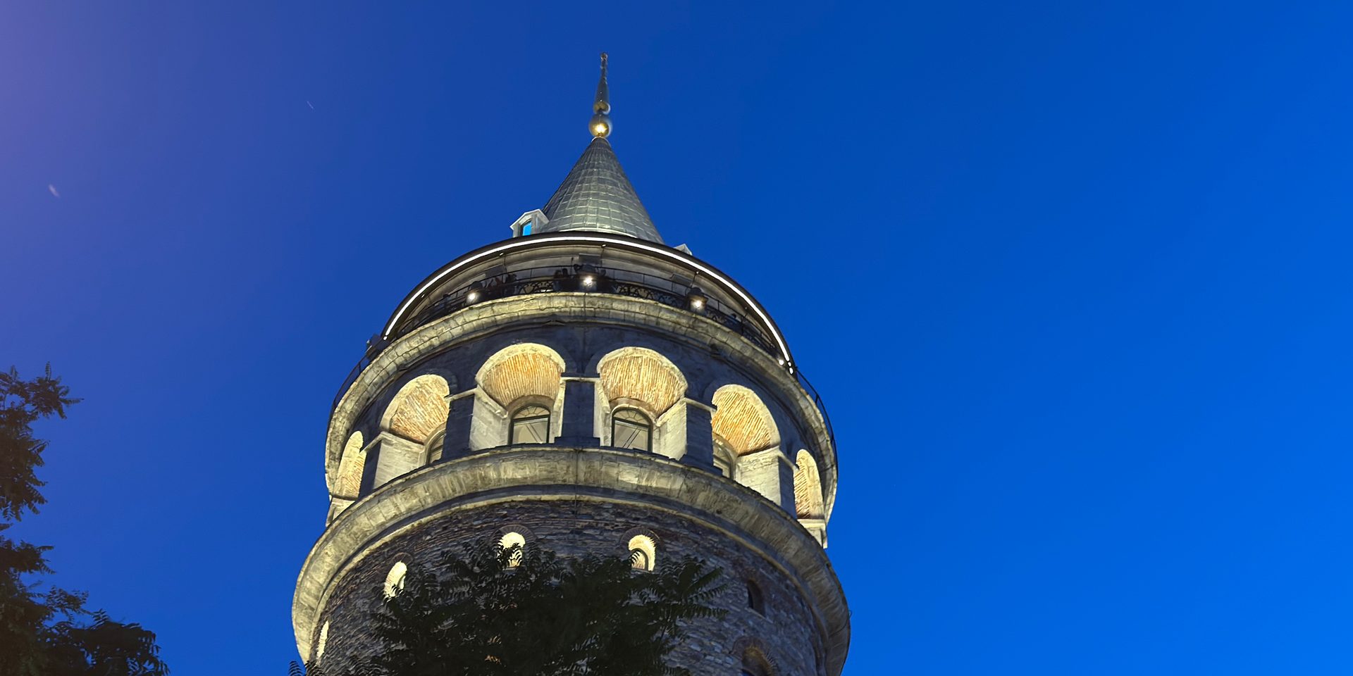 Galata Tower at blue hour: floodlit conical cap, glowing gallery lights, and warm yellow arched windows under a deep blue sky