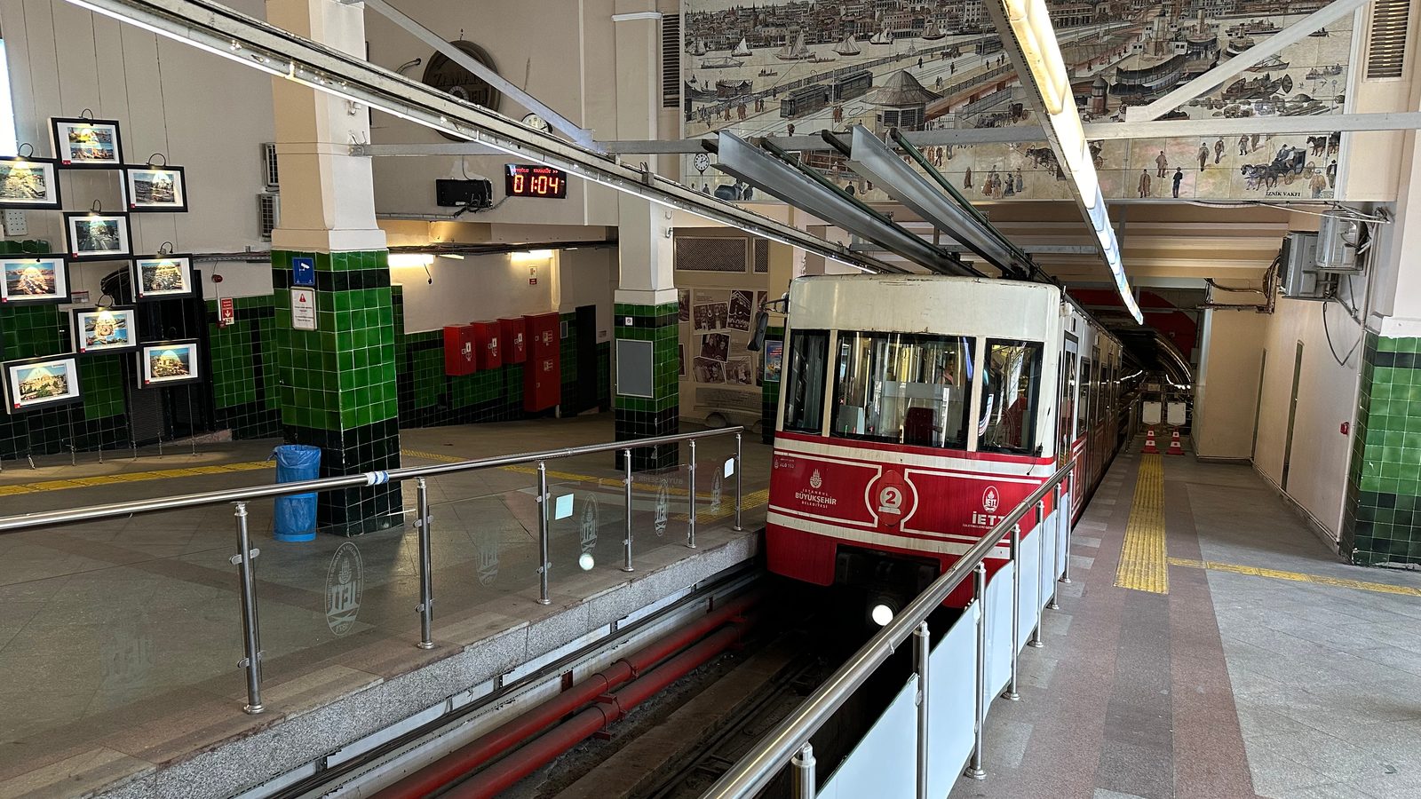Tünel station interior with red-and-cream car on its incline track behind glass railings and green-tiled pillars.