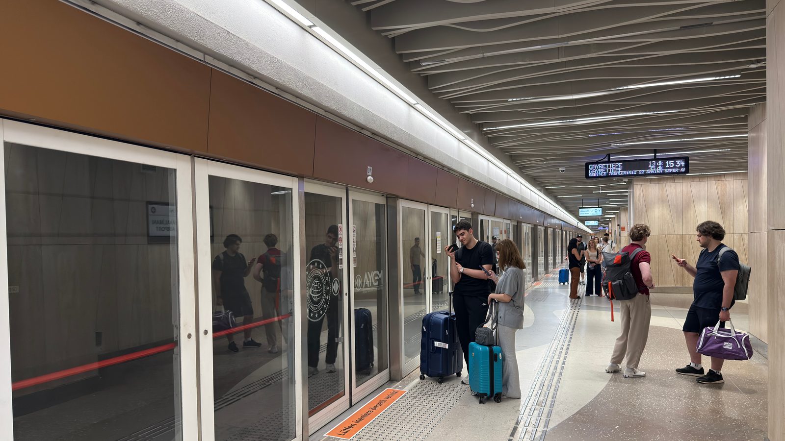 Underground M11 platform at İstanbul Havalimanı with glass screen doors and travellers with suitcases.