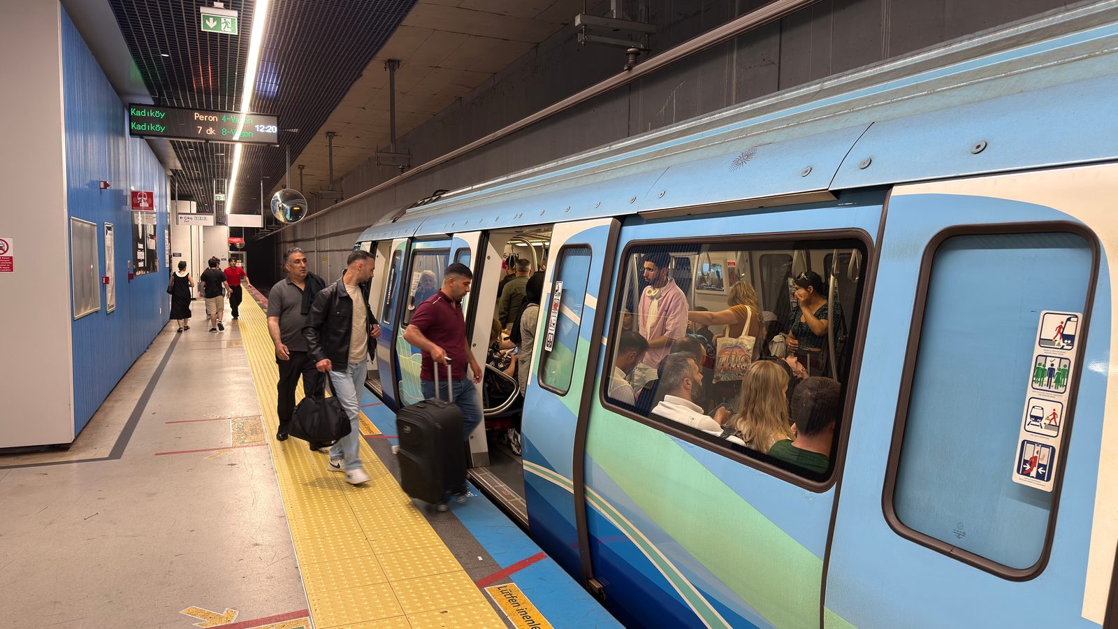 Light-blue and green M4 train with doors open at an underground platform, signs showing Kadıköy.