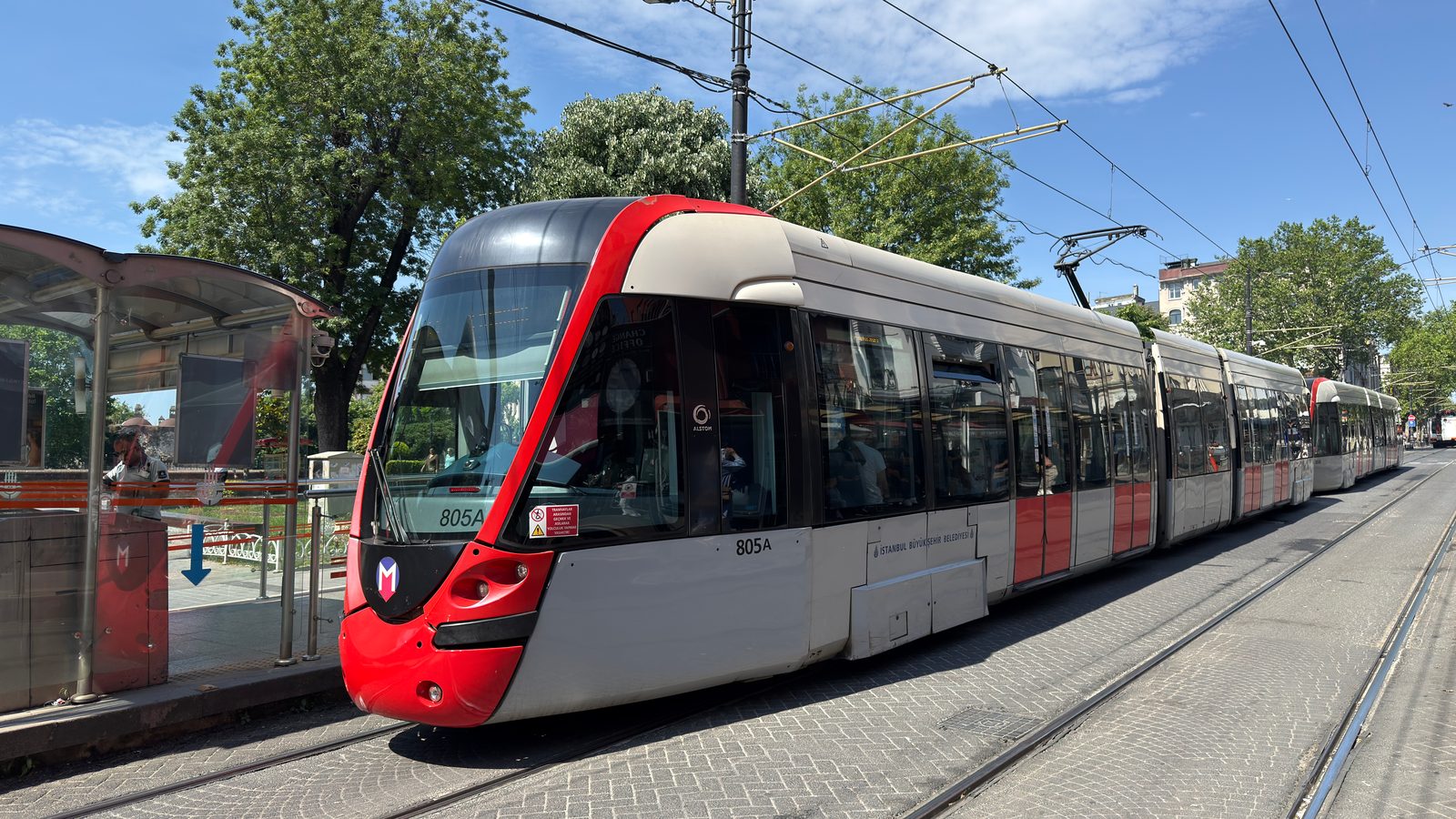 Modern articulated tram at a street stop with shelter, trees and overhead wires.