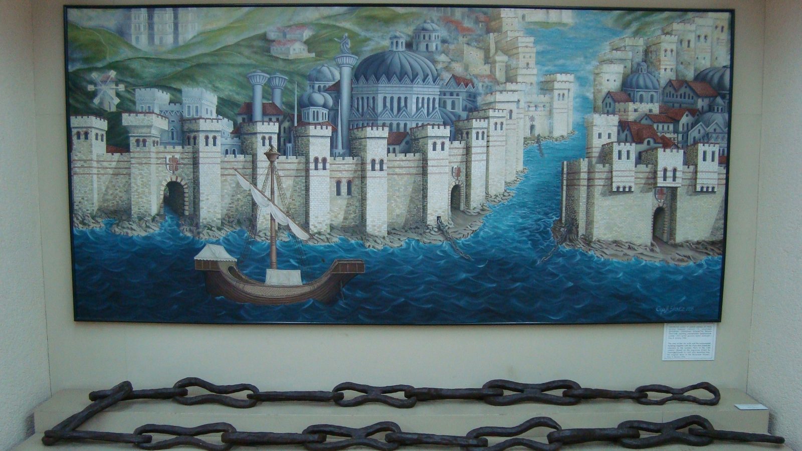 Sections of the massive iron Byzantine chain on a low plinth, displayed beneath a painting of the walled city by the water.