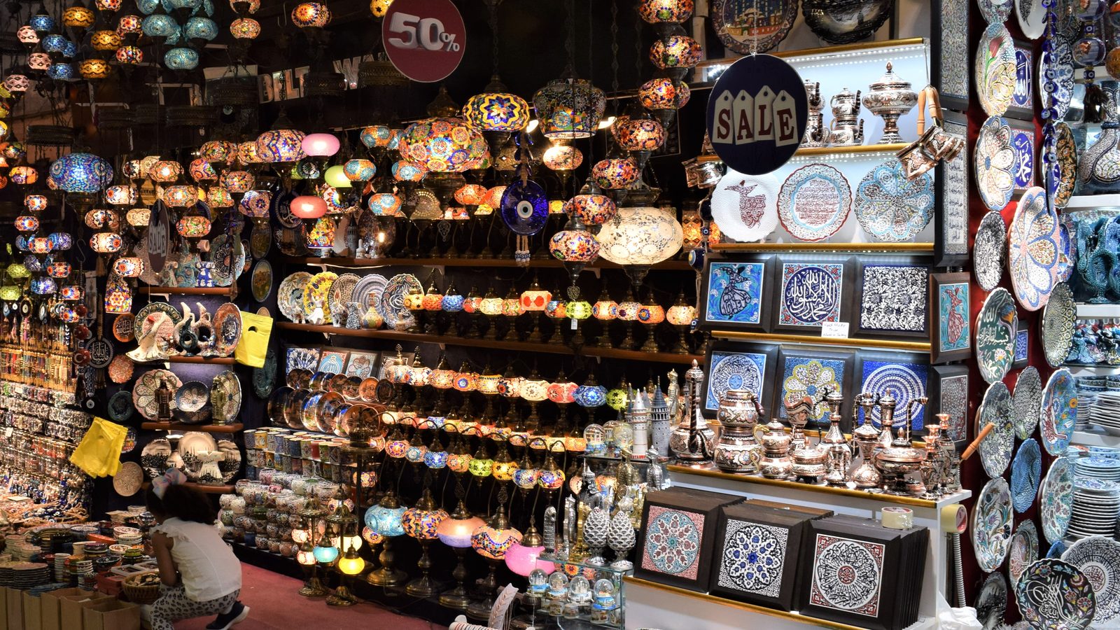 A Grand Bazaar shop front packed with coloured mosaic lamps, ceramic plates and metal teapots, with SALE and 50% signs.