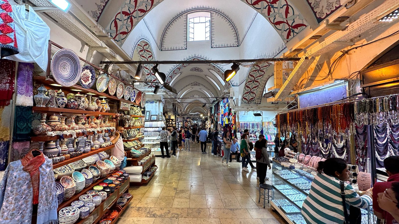 A vaulted Grand Bazaar street lined with ceramics, textile and jewellery shops, shoppers browsing under the arched ceiling.