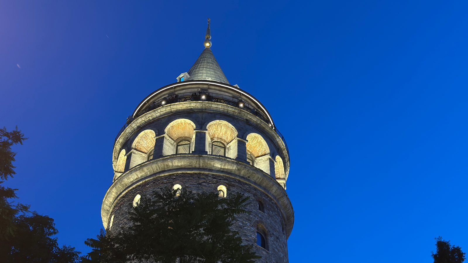 The upper Galata Tower at blue hour under a deep blue sky, its floodlit conical cap, arched windows and observation gallery seen above dark foreground trees