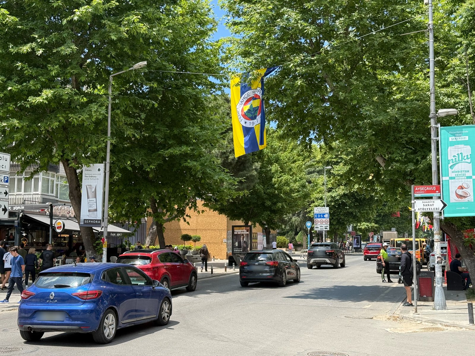 Daytime traffic on Bağdat Caddesi in Suadiye under plane trees, with a Fenerbahçe flag overhead, a traffic officer, and shops.