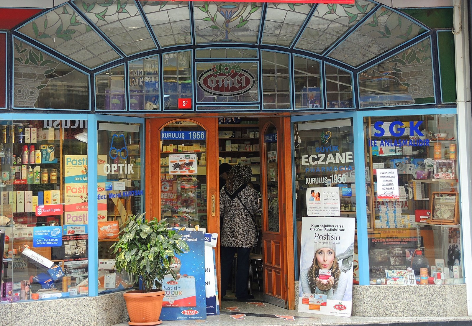 Istanbul pharmacy front with stained-glass canopy, signs for Büyük Eczane and Kuruluş 1956, and a customer in the doorway, back turned.