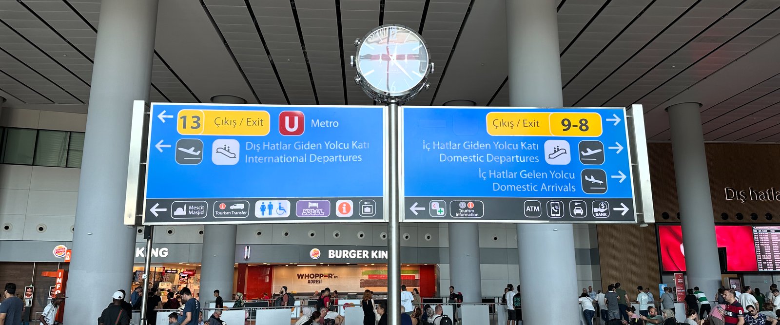Istanbul Airport departures hall: blue overhead signs in Turkish and English for departures, exits, and the metro, with a clock above.