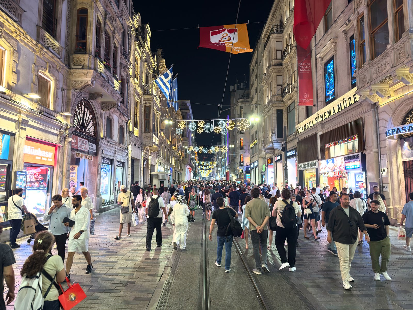 İstiklal Avenue at night: pedestrians on the tram tracks between lit shopfronts, strings of lights overhead, and a sign to the right.