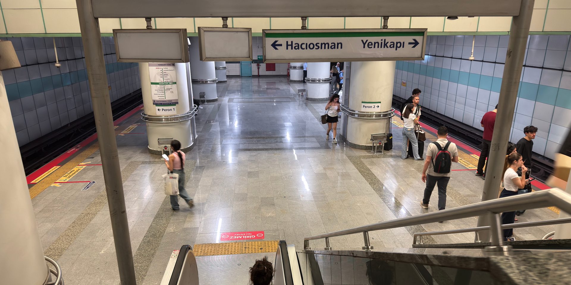 View down an escalator onto an M2 metro platform concourse with Peron 1/2 signs and a few waiting passengers.