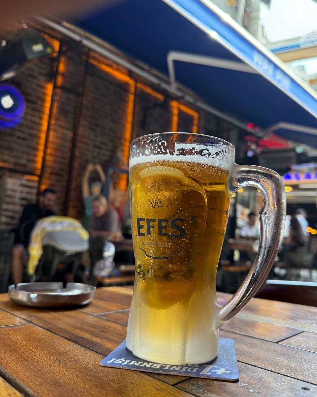 A glass mug of Efes Pilsen beer on a wooden terrace table at dusk, a warm-lit brick wall behind it.